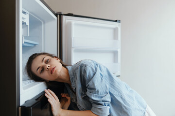 A young woman exhausted and stressed, leaning on an open refrigerator, conveying emotions of...