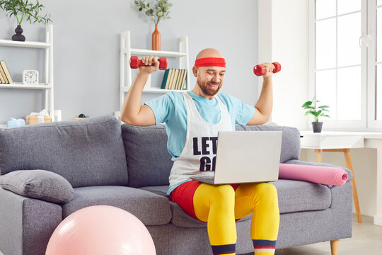 Funny young man in bright sportswear using dumbbells exercising following trainer online via laptop computer sitting on sofa at home. Distant fitness training, sports and humor concept. - Powered by Adobe