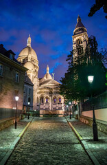 Fototapeta premium Dramatic clouds surround the Roman Catholic Sacré Coeur (Sacred Heart) Basilica, as seen from the north east on Rue du Chevalier de la Barre, at blue hour on a summer evening in Paris, France.