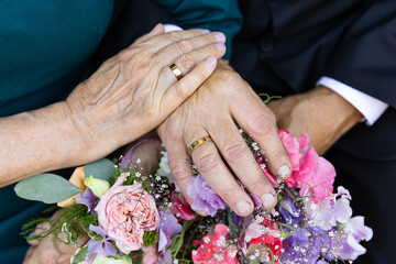 Two hands, worn with age, hold each other gently, showing their wedding rings that have lasted...