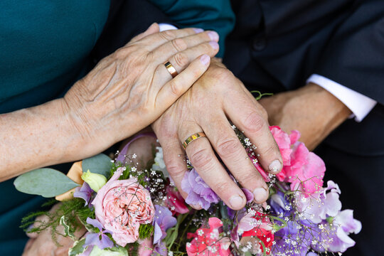 Two hands, worn with age, hold each other gently, showing their wedding rings that have lasted through the years. The rings and flowers tell a story of a lifelong journey together, full of love