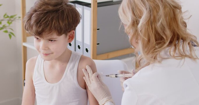 Child gets vaccinated. Boy in a white sleeveless tank top receives an injection at the clinic. Nurse disinfects the skin on the kid's shoulder before the vaccine shot. Health care, vaccination concept