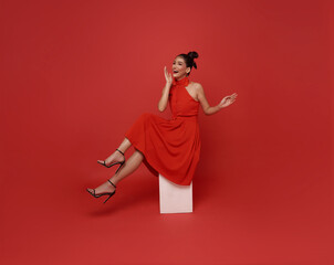 Young beautiful smiling excited asian woman red dress sitting on chair relaxing isolated on red...