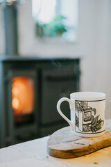 A white mug with a black design sits on a wooden cutting board next to a wood stove. The scene conveys a cozy and warm atmosphere, with the wood stove providing heat