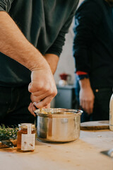 A man is stirring a pot of food on a table. There are two people in the background, one of whom is wearing a watch