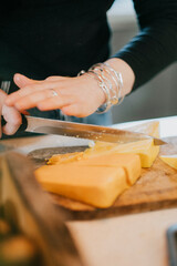 A woman is cutting a piece of cheese on a cutting board. She is wearing a ring and bracelets