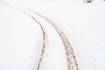A winding dirt road covered in a layer of fresh snow, surrounded by a white landscape. The tire tracks create a contrast against the snow, leading into the distance.