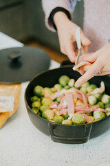 A person is cutting up vegetables in a black pot. The vegetables include broccoli and bacon. The person is using a knife to cut the vegetables