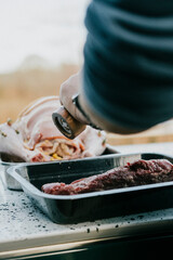 A person is seasoning meat with a pepper grinder. The meat is in a tray and the person is standing next to it