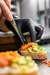 A close-up of a chef's hand in a black glove delicately placing slices of salmon on a colorful bruschetta topped with fresh vegetables and herbs on a dark surface.