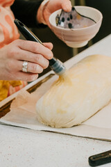 A woman is preparing a pastry by brushing it with a yellow glaze using a black pastry brush. The glaze is spread evenly over the pastry, giving it a glossy and appetizing appearance