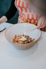 A woman is stirring a bowl of oatmeal with a spoon. The bowl is filled with oatmeal and a small amount of butter. The woman is wearing an apron and she is in the process of preparing breakfast