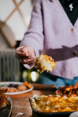 A woman is holding a fork and spoon in front of a plate of food. The food appears to be a type of mashed potato dish. The woman seems to be enjoying her meal and is ready to dig in
