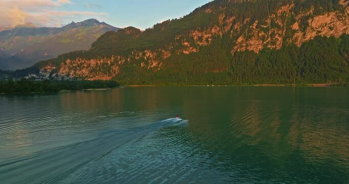 Water skiing dude pulled by the speedboat in the outdoors at blue lake with many luscious, green trees in the background and a cloudy sky, you can see splashes of water or waves. Switzerland