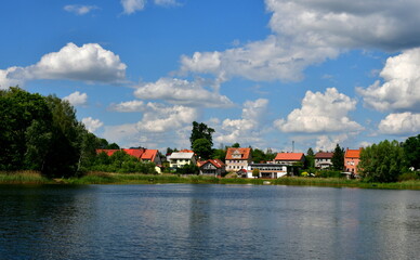 Obraz premium A view of a vast yet shallow river, lake or channel surrounded from all sides with reeds, shrubs and forests with some stone and concrete barriers seen, a small jetty, some boats and a quaint town