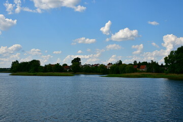 A view of a vast yet shallow river, lake or channel surrounded from all sides with reeds, shrubs and forests with some stone and concrete barriers seen, a small jetty, some boats and a quaint town