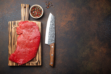 Raw uncooked top round beef steak on wooden cutting board with big kitchen knife and pepper on dark...