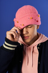 A young man in a pink beanie and hoodie poses, adjusting his hat against a bright backdrop.