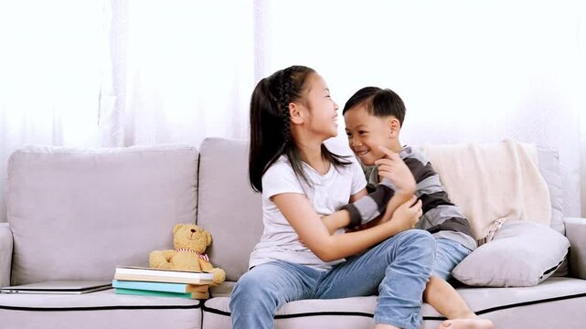 Little brother ran to sister, eager to play with her while she using laptop. Sister playfully pinched brother's cheeks as they sit together on sofa in living room Both siblings look happy laughing 