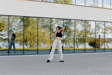 A young woman in a black tank top and white pants dances in front of a large glass building