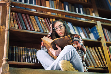 young female student with long hair and glasses sits on a wooden floor with a large old book and reads against the background of bookshelves in the library