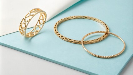 Three Delicate Gold Bangles with Intricate Designs on a Pale Blue Background