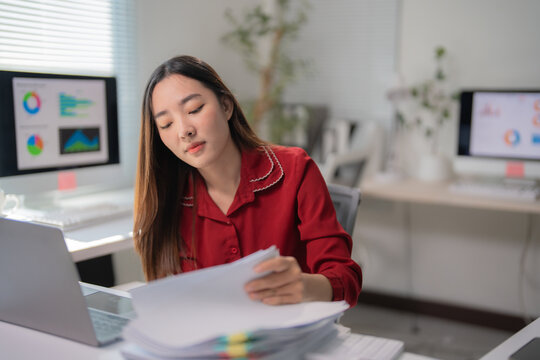 Young asian businesswoman in a red blouse reviewing paperwork while sitting at a desk and working on a laptop in a modern office environment, focused on her tasks and data analysis