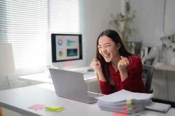 Young happy asian businesswoman raising her arms in front of laptop computer while working in office, celebrating business success, achievement and expressing excitement