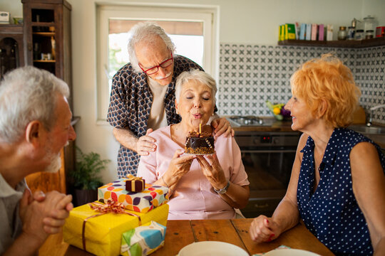 Group of senior friends celebrating birthday blowing cake candle at kitchen table