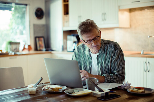 Worried senior man reading paperwork while working on laptop at kitchen desk