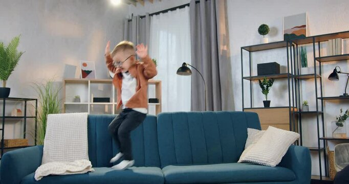 Little boy having fun alone while jumping on the soft couch in contemporary apartment