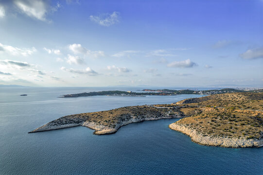 A rocky coastline with small islands and boats in the distance, surrounded by a calm blue sea and a partly cloudy sky. Limanakia beach, Vouliagmeni, Athens