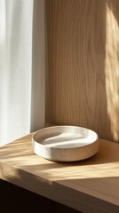A simple, round ceramic dish rests on a wooden shelf, illuminated by soft natural light.
