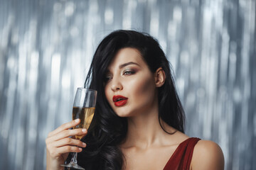 Brunette woman in a red dress holding a champagne glass with a relaxed and slightly tipsy expression, set against a glittering silver backdrop. Great for lifestyle, party, or festive themes. 