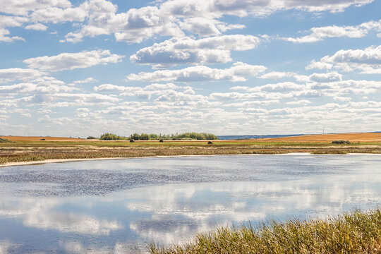 Cloudscape with clouds reflected in water with hay bale field in the distance, a typical rural Saskatchewan scene.