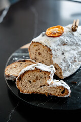 A festive Christmas Stollen covered in powdered sugar, garnished with dried orange and cinnamon, showing sliced portions revealing dried fruit and nuts. Cozy holiday atmosphere  .