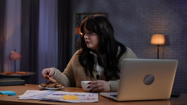 Young business woman freelancer sitting at desk working on remote in home office late, using laptop, drinking coffee wants to eat cookie, doubting and refuses.