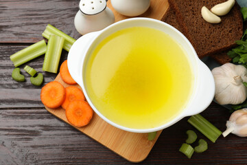 Tasty chicken bouillon in bowl with bread and ingredients on wooden table, flat lay