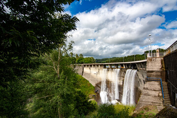 The image shows a large concrete dam with water cascading down its face from multiple spillways. The structure is massive and surrounded by lush green vegetation and cloudy skies.