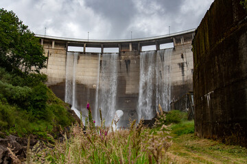 The image shows a large concrete dam with water cascading down its face from multiple spillways. The structure is massive and surrounded by lush green vegetation and cloudy skies.