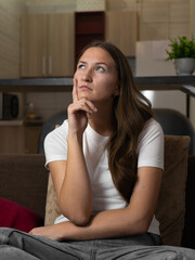 Young woman is sitting on a sofa at home, touching her chin with her hand and looking up...