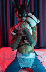 Sexy guy in bdsm rabbit mask and leather belt on a bed in neon light waiting for sex with a mistress with a toy