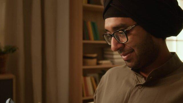 Tilt shot of happy Indian man with stubble, wearing glasses, traditional kurta and turban, texting messages on smartphone to beloved girlfriend and smiling, resting at home