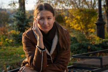 Young woman suffering from headache while sitting on bench in park during autumn