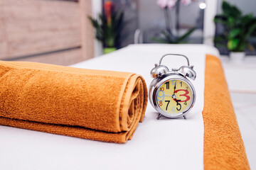 A cozy spa setup featuring an orange rolled towel and a retro alarm clock on a massage table. Ideal for relaxation, wellness, and time management themes.