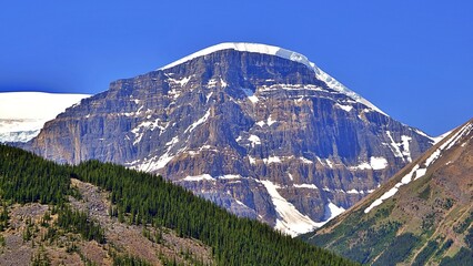 A stunning photograph of a snow-capped mountain peak rising sharply against a clear blue sky. The dramatic rock formations and patches of snow create a striking contrast, showcasing the rugged beauty 