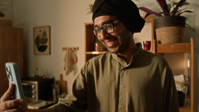 Tilt shot of happy smiling Indian man in glasses, traditional khaki kurta and black head hugger talking during video call on smartphone and waving in good-bye at home, ending online conversation