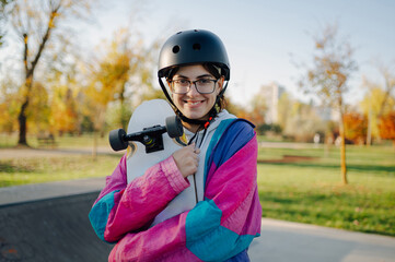 Young skater holding her skateboard smiling in a park during a sunny autumn day