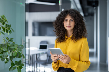 A professional woman with curly hair, uses a digital tablet in a modern office setting. The...