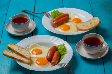 Breakfast fried eggs with sausages crispy white bread and greens, and tea on a blue table